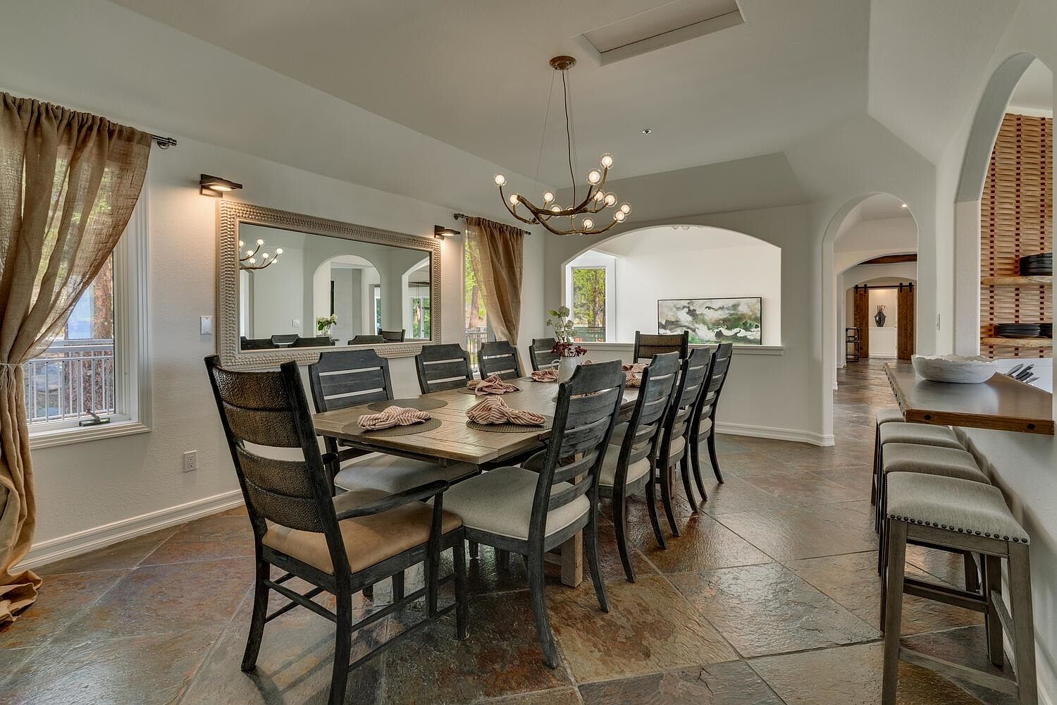 This is an interior shot of a dining room featuring a long wooden table with seating for ten. The room has a large mirror on one wall, reflecting the table and chairs, and a modern chandelier hangs above the table. The flooring is tile, and there are arched doorways leading to other parts of the house, creating an open and inviting space.
