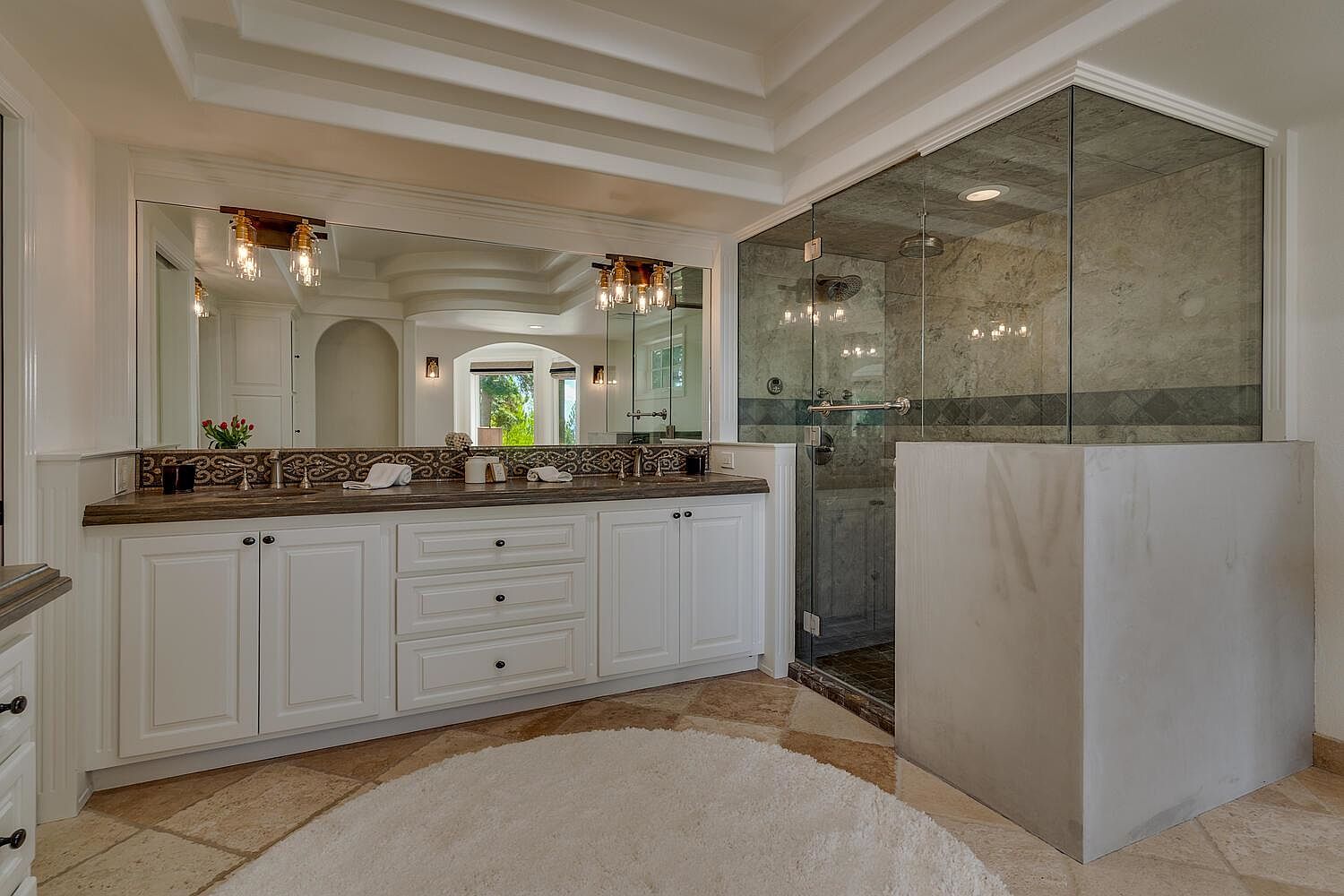 This is a primary bathroom featuring a double vanity with white cabinetry and a dark countertop, complemented by a large mirror and decorative lighting fixtures. A glass-enclosed shower with marble-like tiling is visible to the right, adding a luxurious touch. The flooring is tiled in a warm tone, and a round white rug adds softness to the space.