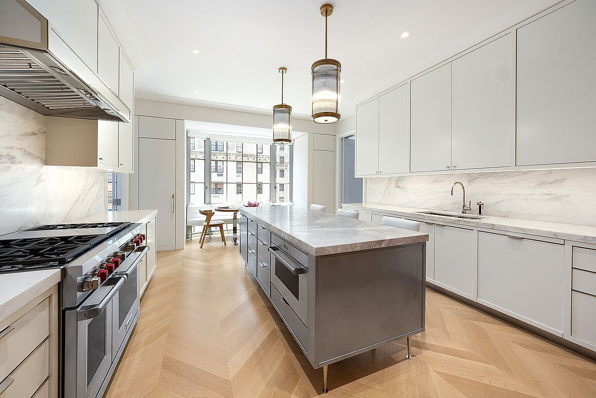 This sophisticated, high-end kitchen features a central island with a stone countertop and a contrasting dark base, complemented by light-toned cabinetry and matching stone backsplashes. The space is illuminated by elegant pendant lighting and includes professional-grade stainless steel appliances, including a large range and hood. The room is finished with light wood flooring laid in a chevron pattern, creating a bright and airy atmosphere that flows toward a sunlit breakfast area.