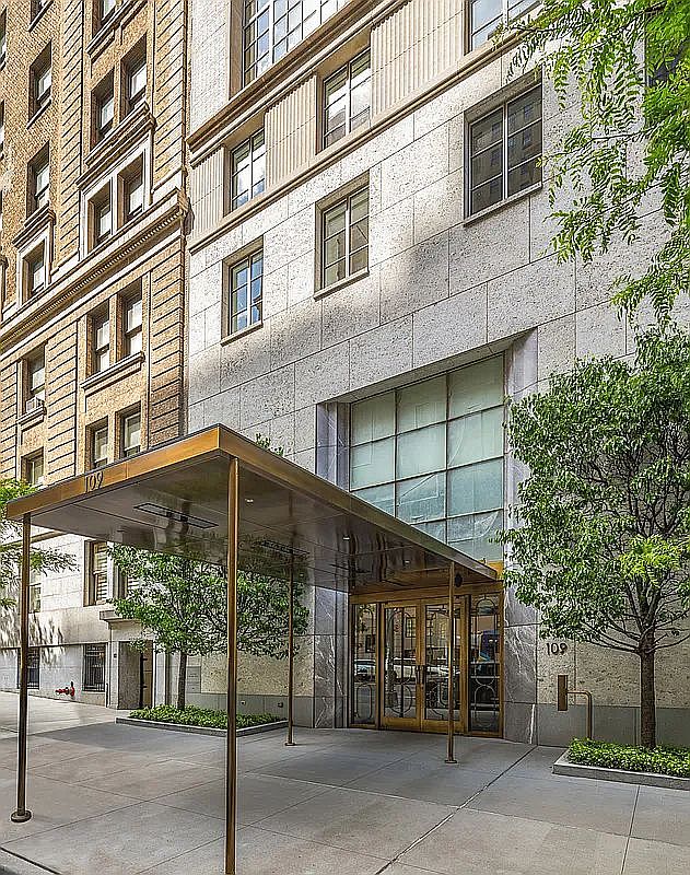 This image captures the sophisticated street-level entrance of a multi-story building, featuring a prominent metal canopy extending over the sidewalk. The facade is composed of light-colored stone panels, complemented by a large glass window wall above the main entrance doors. A small, manicured tree stands to the right, adding a touch of greenery to the urban architectural setting.