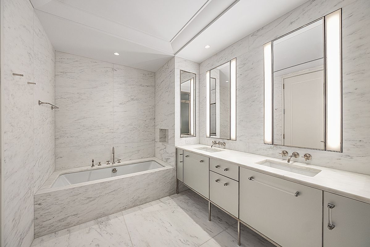 This luxurious primary bathroom features extensive light-toned stone surfaces throughout the walls and flooring, creating a bright and cohesive aesthetic. The space includes a deep soaking tub set against a stone-clad wall, alongside a long vanity with dual sinks and integrated vertical lighting flanking the mirrors. The clean lines and minimalist hardware contribute to a sophisticated, high-end atmosphere.