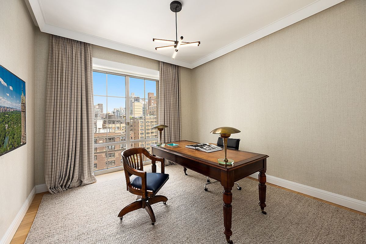 This sophisticated home office features a classic wooden desk centered in the room, paired with a traditional swivel chair and a sleek modern task chair. The space is defined by neutral textured wallpaper, a large area rug, and tall windows that provide an expansive urban view. A contemporary multi-arm chandelier hangs from the ceiling, adding a touch of modern elegance to the professional setting.