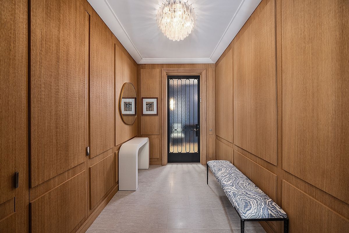This elegant hallway features warm wood-paneled walls with classic molding, creating a sophisticated and cohesive aesthetic. A central chandelier provides soft illumination, while a sleek, curved console table and a patterned bench offer functional and stylish accents. The perspective looks directly down the corridor toward a dark-framed glass door, emphasizing the symmetrical and refined design of the space.