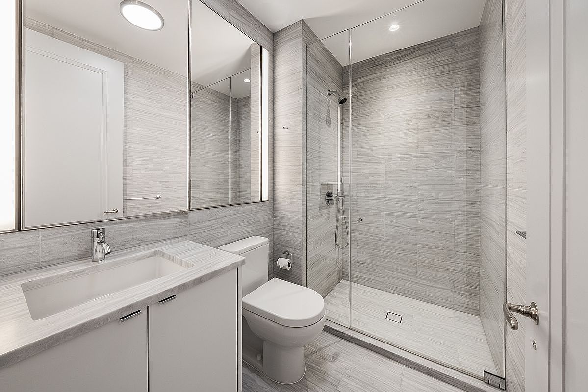 This modern, compact bathroom features a sleek vanity with a stone countertop and a large, integrated mirror cabinet. The space is finished with light-toned, horizontal-patterned wall tiles that extend into the walk-in shower, which is enclosed by a clear glass panel. A white toilet sits between the vanity and the shower, creating a clean and efficient layout.