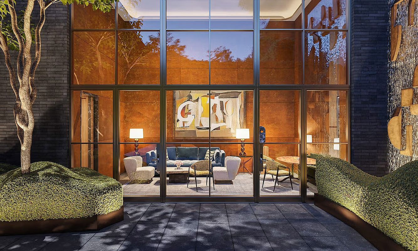 This image captures a sophisticated lounge area viewed from an outdoor courtyard, framed by expansive floor-to-ceiling glass walls. The interior features a plush blue sofa, modern armchairs, and a large abstract painting, creating a refined and inviting atmosphere. The exterior space is landscaped with manicured hedges and a tree, set against dark brick walls, offering a seamless transition between indoor and outdoor living.