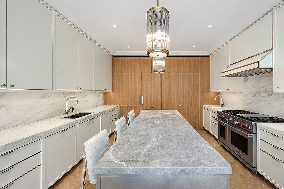 This sophisticated, high-end kitchen features a central island with a large stone countertop and integrated seating, flanked by sleek white cabinetry and light wood paneling. The space is illuminated by a striking tiered pendant light fixture, while stone backsplashes and professional-grade appliances add a polished, functional aesthetic. The perspective is a centered, eye-level shot that emphasizes the symmetrical layout and clean, modern lines of the room.