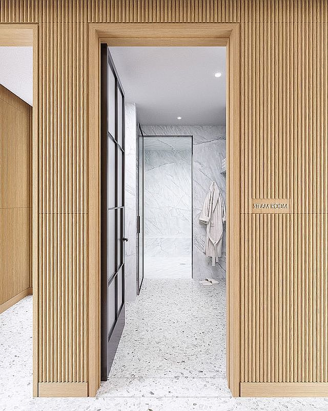 This image captures the entrance to a steam room, characterized by elegant, light-toned vertical wood paneling that creates a sophisticated, textured aesthetic. A dark-framed glass door leads into a bright, minimalist space featuring light-colored stone flooring and walls, with a bathrobe hanging on the wall. The perspective is centered on the doorway, emphasizing the clean lines and high-end, spa-like atmosphere of the facility.