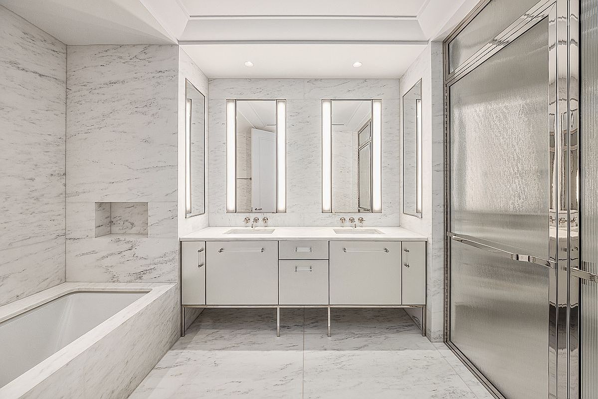 This luxurious primary bathroom features a double vanity with a light-toned cabinet base, topped with a stone counter and dual sinks. The space is clad in light-colored stone with subtle veining, including a built-in bathtub area and a glass-enclosed shower stall. The symmetrical layout is enhanced by vertical wall-mounted lighting fixtures flanking the mirrors, creating a bright and sophisticated atmosphere.