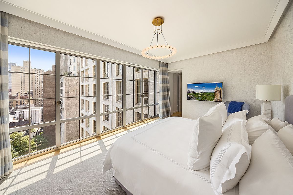 This bright and airy primary bedroom features a large, plush bed centered in the room, complemented by a modern circular chandelier overhead. A wall of expansive windows provides abundant natural light and a clear view of the surrounding city architecture. The space is finished with neutral-toned walls and soft carpeting, creating a serene and sophisticated atmosphere.