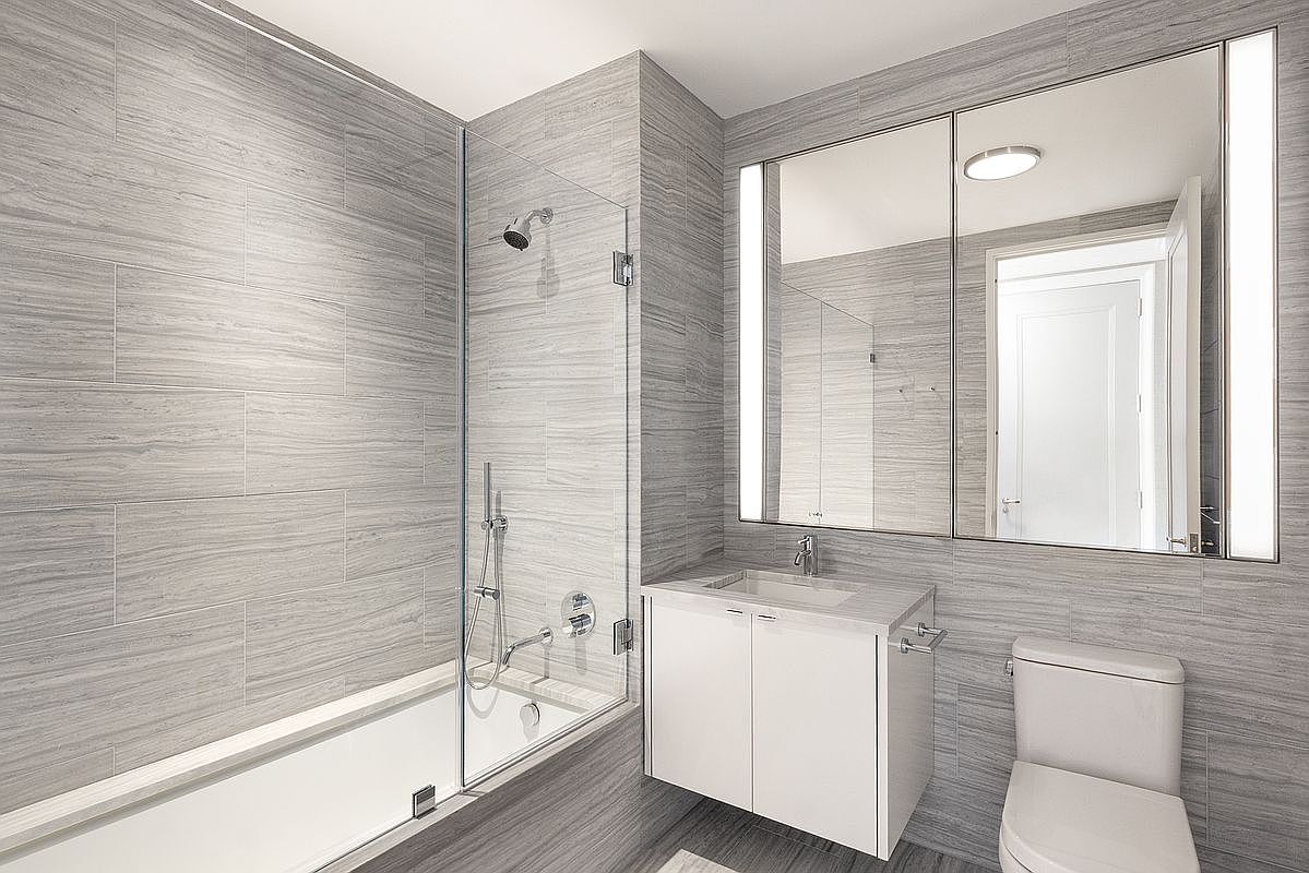This modern bathroom features a sleek, minimalist design with grey wood-look tile covering the walls and floor. The space includes a white vanity with a stone countertop, a glass-enclosed shower-tub combination, and a large, integrated mirror with vertical light strips. The perspective is a wide-angle shot capturing the entire functional layout of the room, emphasizing its clean lines and contemporary aesthetic.
