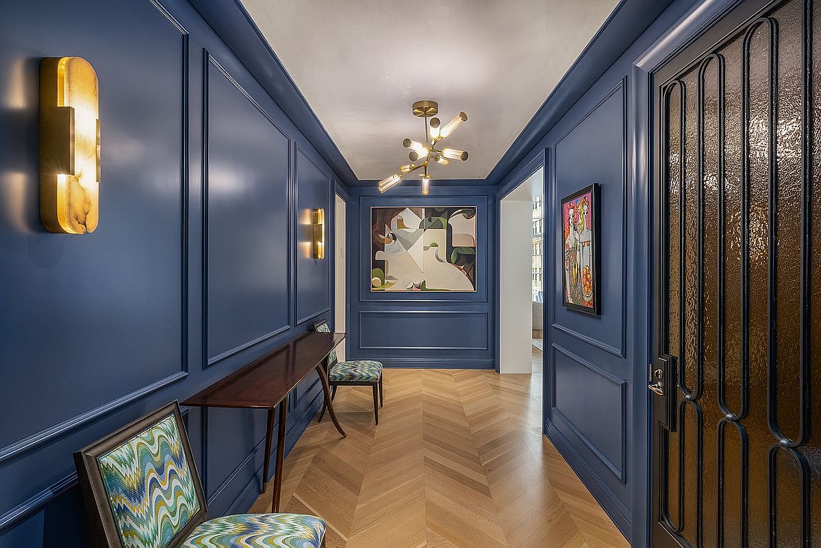 This sophisticated hallway features deep blue paneled walls and a light-toned wood floor laid in a chevron pattern. The space is accented by modern gold-toned light fixtures, including a sculptural chandelier and wall sconces, alongside colorful abstract artwork. A slim console table and patterned chairs provide functional elegance, creating a refined and welcoming transition space.