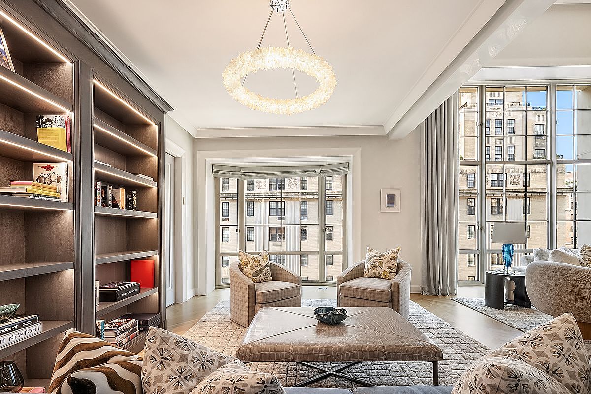This sophisticated living room features a large, textured ottoman at its center, flanked by two patterned armchairs and a built-in bookshelf with integrated lighting. The space is illuminated by a striking, circular chandelier and benefits from abundant natural light streaming through large windows that offer an urban view. The neutral color palette and plush furnishings create an elegant and inviting atmosphere suitable for a high-end residence.