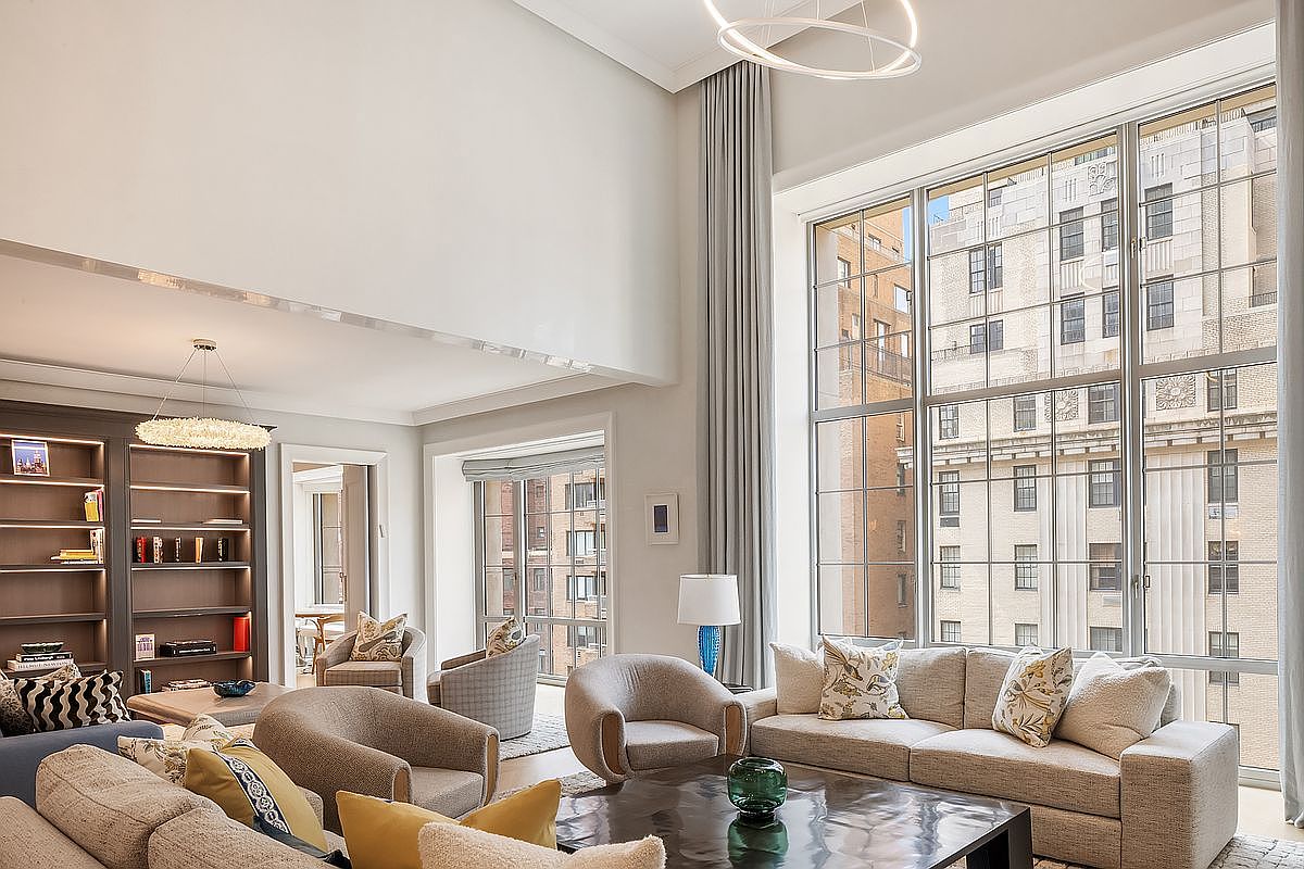 This expansive, high-ceilinged living room features a sophisticated neutral color palette and a grand wall of tall windows that flood the space with natural light. The room is furnished with plush, contemporary seating, including a large sofa and several armchairs arranged around a dark, reflective coffee table. A built-in dark wood bookshelf adds a touch of contrast, while elegant light fixtures and soft drapery enhance the refined, airy atmosphere of this luxury residence.