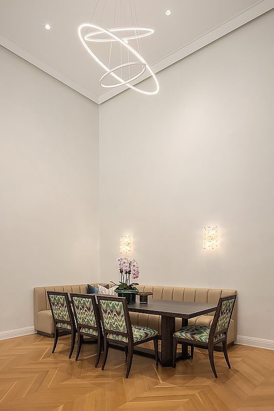 This elegant dining area features a built-in corner banquette paired with a dark wood table and four chairs upholstered in a vibrant, multi-colored chevron pattern. The space is defined by high ceilings, light-toned walls, and warm-toned wood flooring arranged in a herringbone pattern. A modern, sculptural ring chandelier hangs above the table, complemented by two matching wall sconces that provide soft, ambient lighting.