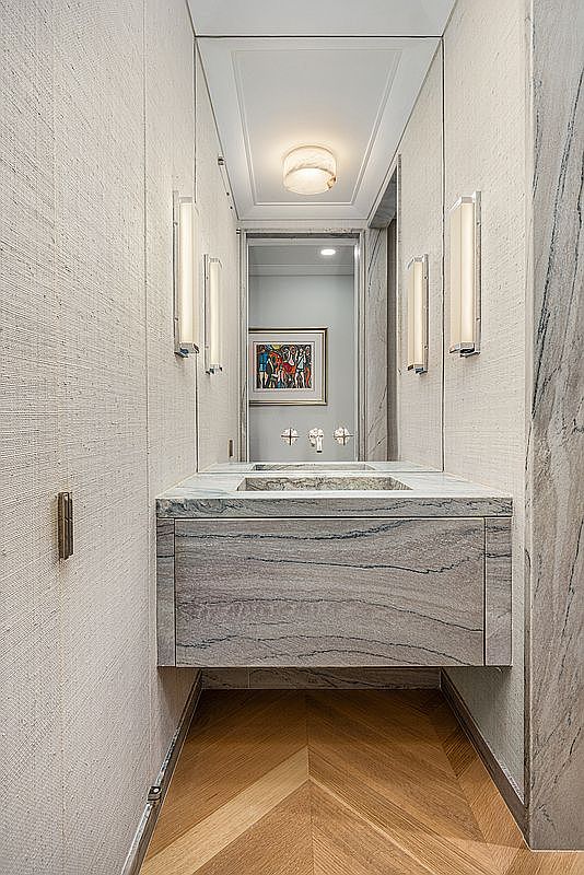 This elegant powder room features a floating stone vanity with prominent veining, set against textured wall coverings. The space is framed by a large mirror that reflects a framed piece of art in the adjacent room, while vertical wall-mounted light fixtures provide soft illumination. The floor is finished in a light-toned wood arranged in a classic chevron pattern, creating a sophisticated and cohesive aesthetic.