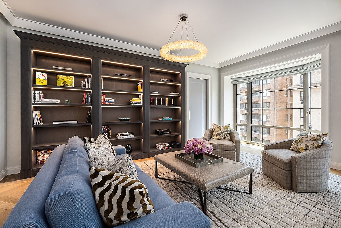 This sophisticated living room features a large, dark-toned built-in bookshelf with integrated LED lighting that spans the entire wall. A plush blue sofa with patterned throw pillows sits opposite two comfortable armchairs, all centered around a large, textured ottoman coffee table topped with a floral arrangement. The room is bathed in natural light from a large window wall, creating an inviting and elegant atmosphere.
