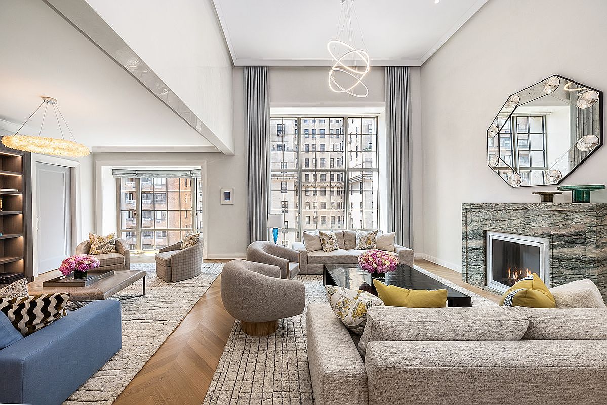 This expansive, high-ceilinged living room features a sophisticated, neutral color palette with layered textures and herringbone wood flooring. A prominent stone-clad chimney breast serves as a focal point, complemented by a large, multi-faceted decorative mirror and a contemporary light fixture. The space is bathed in natural light from tall windows that offer an urban view, creating an airy and luxurious atmosphere.