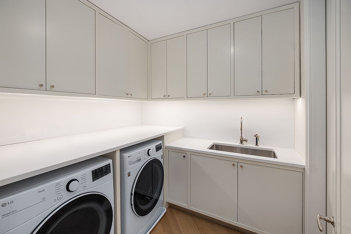 This clean and modern laundry room features light-toned cabinetry providing ample storage space above and below a white countertop. The room is equipped with side-by-side LG appliances and a built-in stainless steel sink with a sleek faucet. Under-cabinet lighting illuminates the workspace, creating a bright and functional environment.