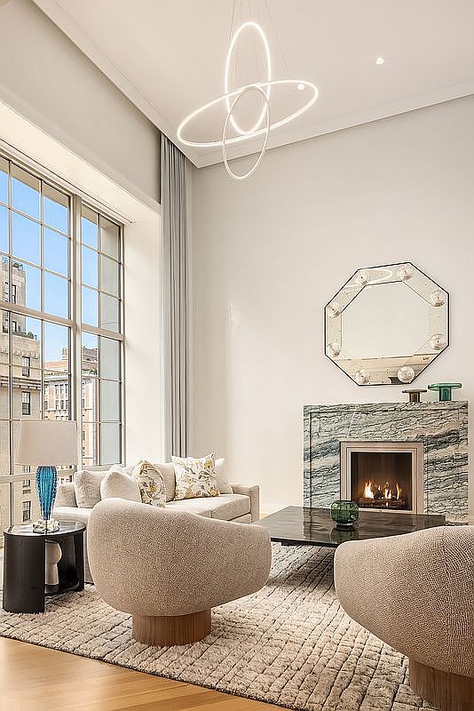 This sophisticated living room features a striking, modern sculptural chandelier suspended from a high ceiling, casting a refined glow over the space. A prominent fireplace with a dramatic, veined stone surround serves as the focal point, complemented by an octagonal mirror and plush, curved seating arranged on a textured area rug. Large, multi-paned windows allow ample natural light to fill the room, offering a view of the urban architecture outside.