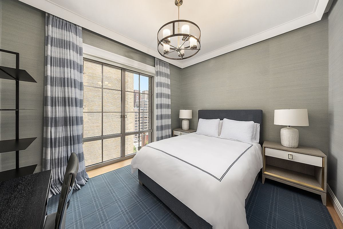 This well-appointed bedroom features a neutral color palette with textured gray walls and a blue plaid area rug. A queen-sized bed with a dark upholstered headboard is centered between two matching nightstands topped with white table lamps. Large windows with blue and white striped curtains allow natural light to fill the space, while a modern circular chandelier hangs from the ceiling.