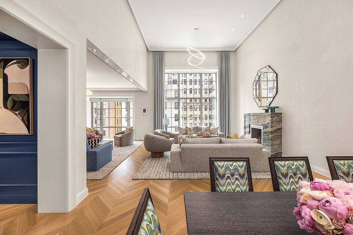 This sophisticated living area features a high-ceilinged layout with herringbone wood flooring and a neutral color palette. A prominent stone-clad chimney breast serves as a focal point, complemented by a large, modern chandelier and expansive windows that flood the space with natural light. The room flows seamlessly into an adjacent seating area, creating an open and airy atmosphere ideal for entertaining.