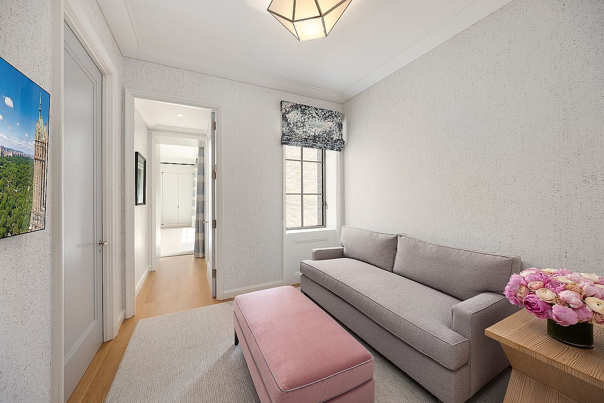 This cozy sitting area features a light gray sofa paired with a soft pink ottoman, creating a refined and inviting atmosphere. The room is accented by textured wallpaper, a geometric pendant light fixture, and a window dressed in a patterned Roman shade. A doorway leads into a bright, neutral-toned hallway, providing a sense of depth and flow to the home's layout.