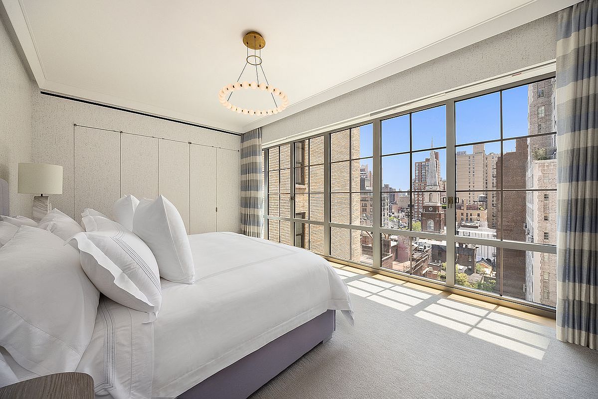 This bright and airy primary bedroom features a plush bed dressed in white linens, positioned beside a wall of large windows that offer an expansive urban view. The room is accented by a modern, circular chandelier and soft, neutral-toned carpeting, creating a serene and sophisticated atmosphere. The clean lines and minimalist aesthetic are complemented by floor-to-ceiling drapery and built-in closet doors along the adjacent wall.