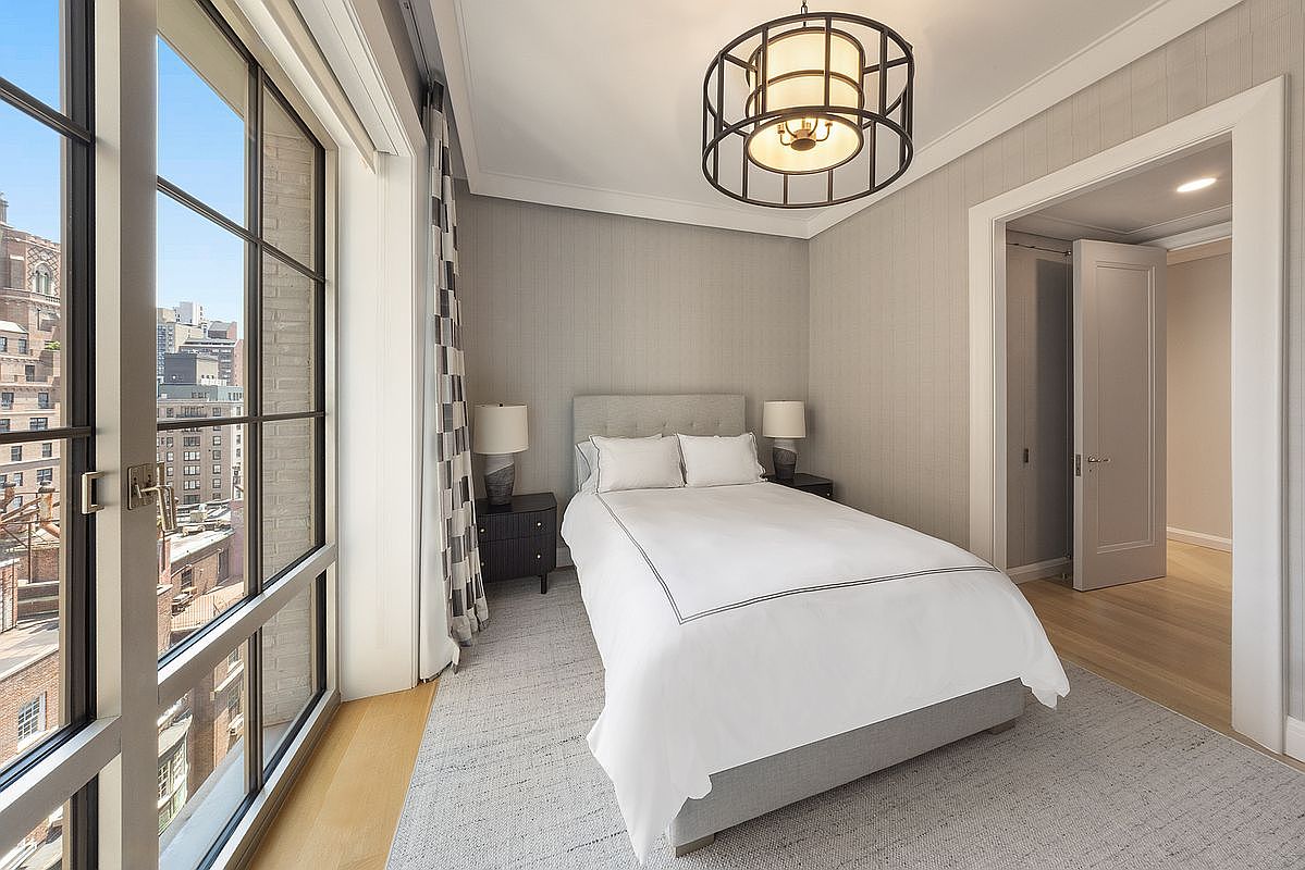 This elegant bedroom features a neutral color palette with textured wallpaper and a plush, light-toned area rug. A central bed with crisp white linens is flanked by two dark wood nightstands and matching lamps, all illuminated by a distinctive geometric pendant light. Large windows with a grid pattern provide an expansive view of the surrounding city architecture, creating a bright and sophisticated urban retreat.