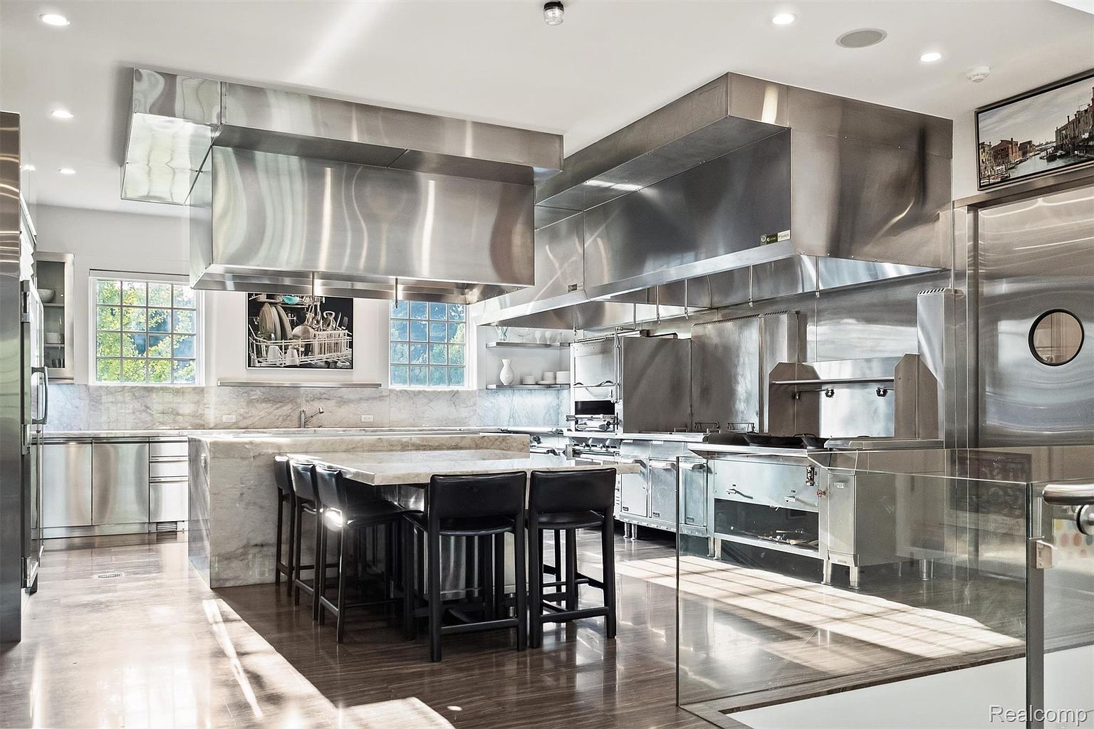 This is a professional-grade kitchen featuring stainless steel appliances and fixtures, including large ventilation hoods and a central island with seating. The design emphasizes functionality and durability, with marble countertops and a sleek, modern aesthetic. Natural light floods the space, highlighting the clean lines and reflective surfaces.