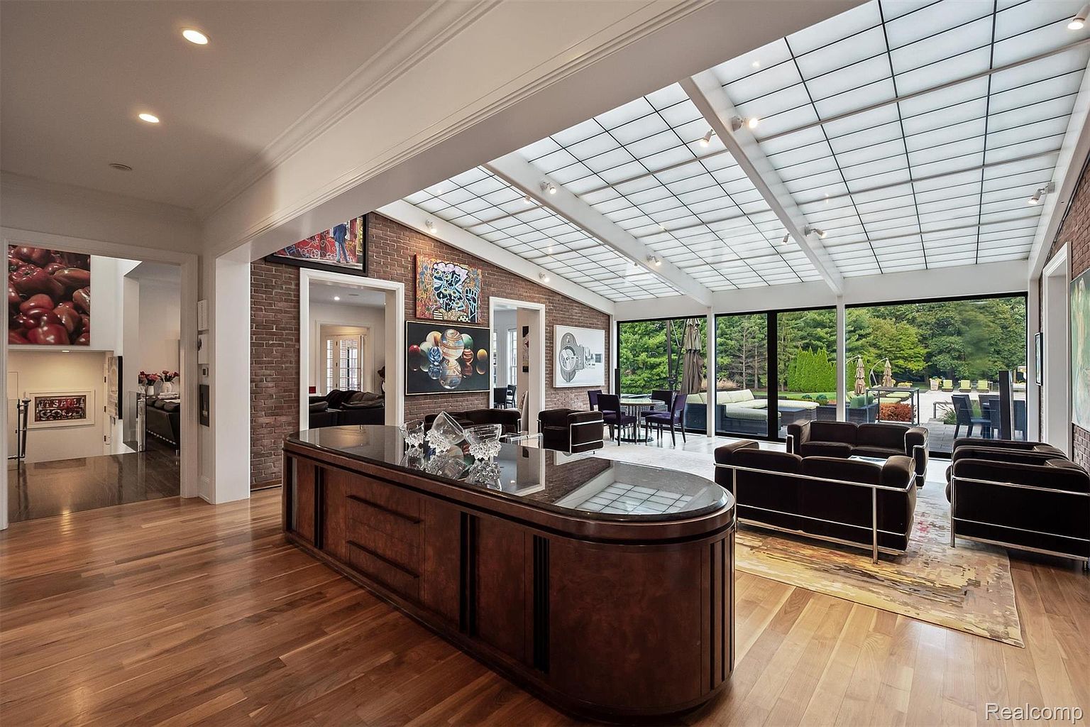 This is an interior shot of a spacious living room featuring hardwood floors, a brick accent wall adorned with artwork, and a unique glass-paneled ceiling that floods the room with natural light. The room is furnished with modern sofas and chairs, and large windows offer views of the outdoor landscape, creating a bright and inviting atmosphere. A large, dark wood cabinet with a glossy top serves as a focal point in the foreground.