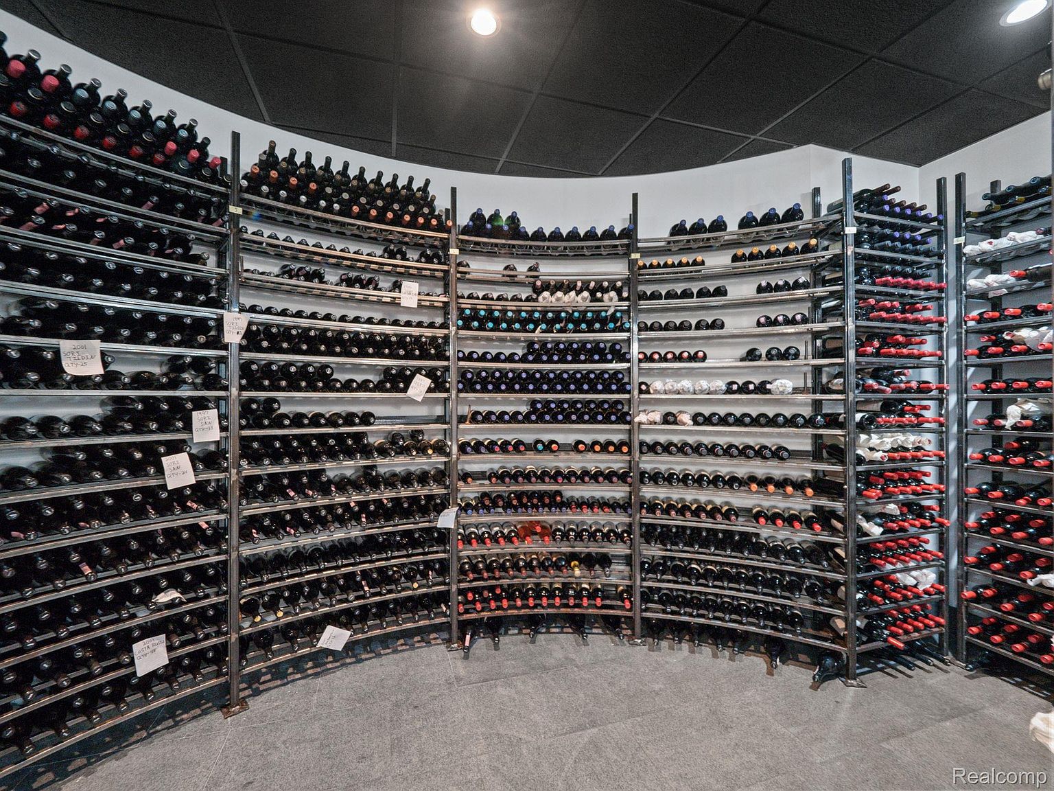 This image showcases a well-stocked wine cellar with curved metal racks filled with numerous bottles of wine. The racks are arranged in a semi-circular fashion, creating an organized and visually appealing display. The cellar features a dark ceiling and a gray tiled floor, contributing to a cool and controlled environment ideal for wine storage.