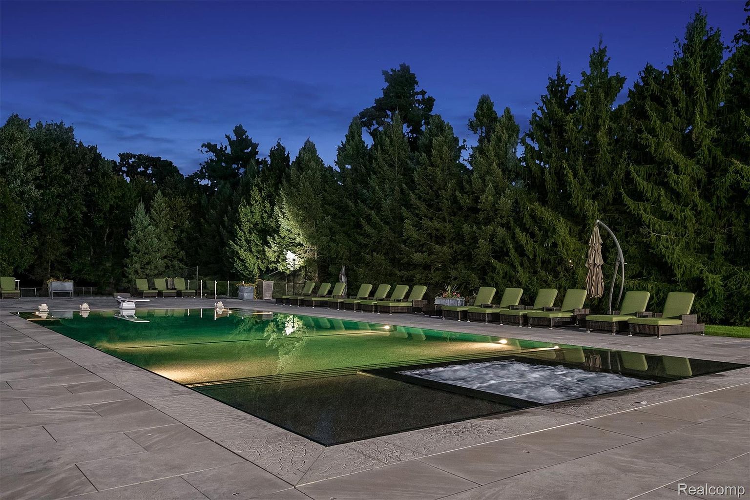 This image showcases a luxurious outdoor pool and spa area at dusk. The rectangular pool features a dark bottom and is surrounded by a stone patio, with lounge chairs lined up along one side. Lush greenery and trees provide a serene backdrop, enhancing the property's appeal for relaxation and entertainment.
