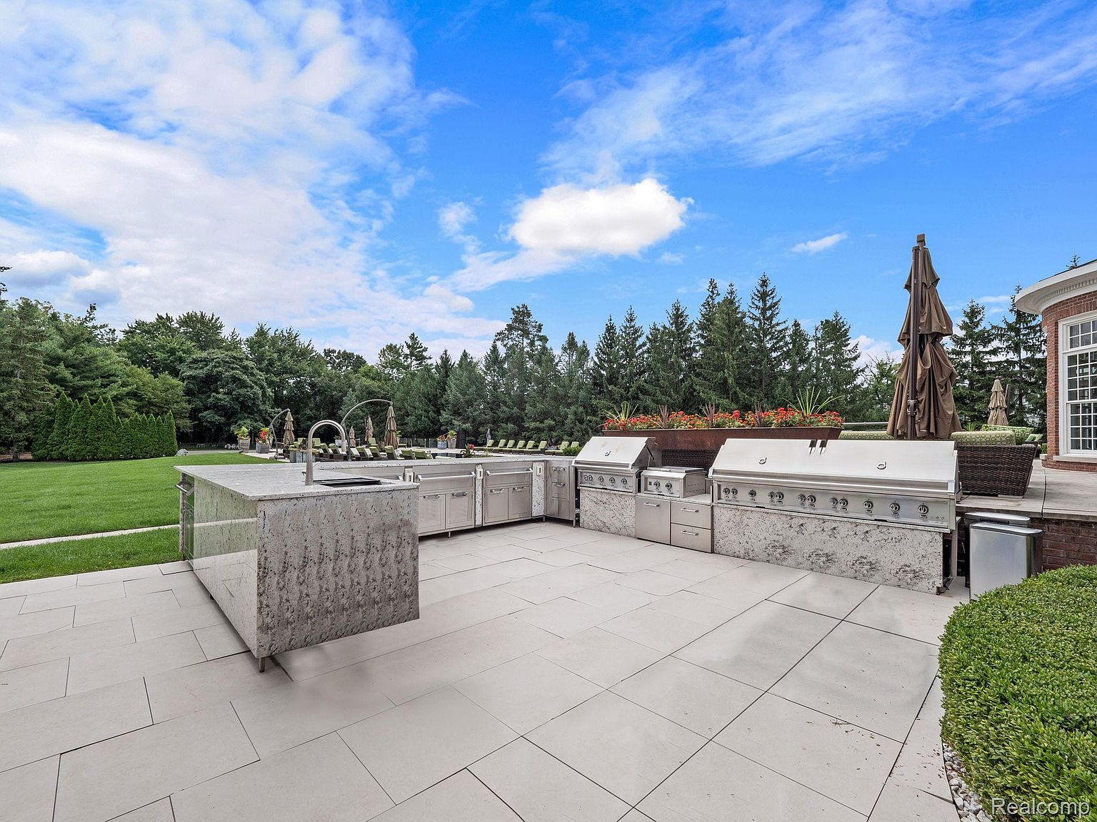 This image showcases a luxurious outdoor kitchen and patio area. The space features high-end stainless steel appliances, including multiple grills and storage units, set against a backdrop of lush greenery and a clear blue sky. The patio is paved with large, light-colored tiles, creating a clean and modern aesthetic, perfect for entertaining.
