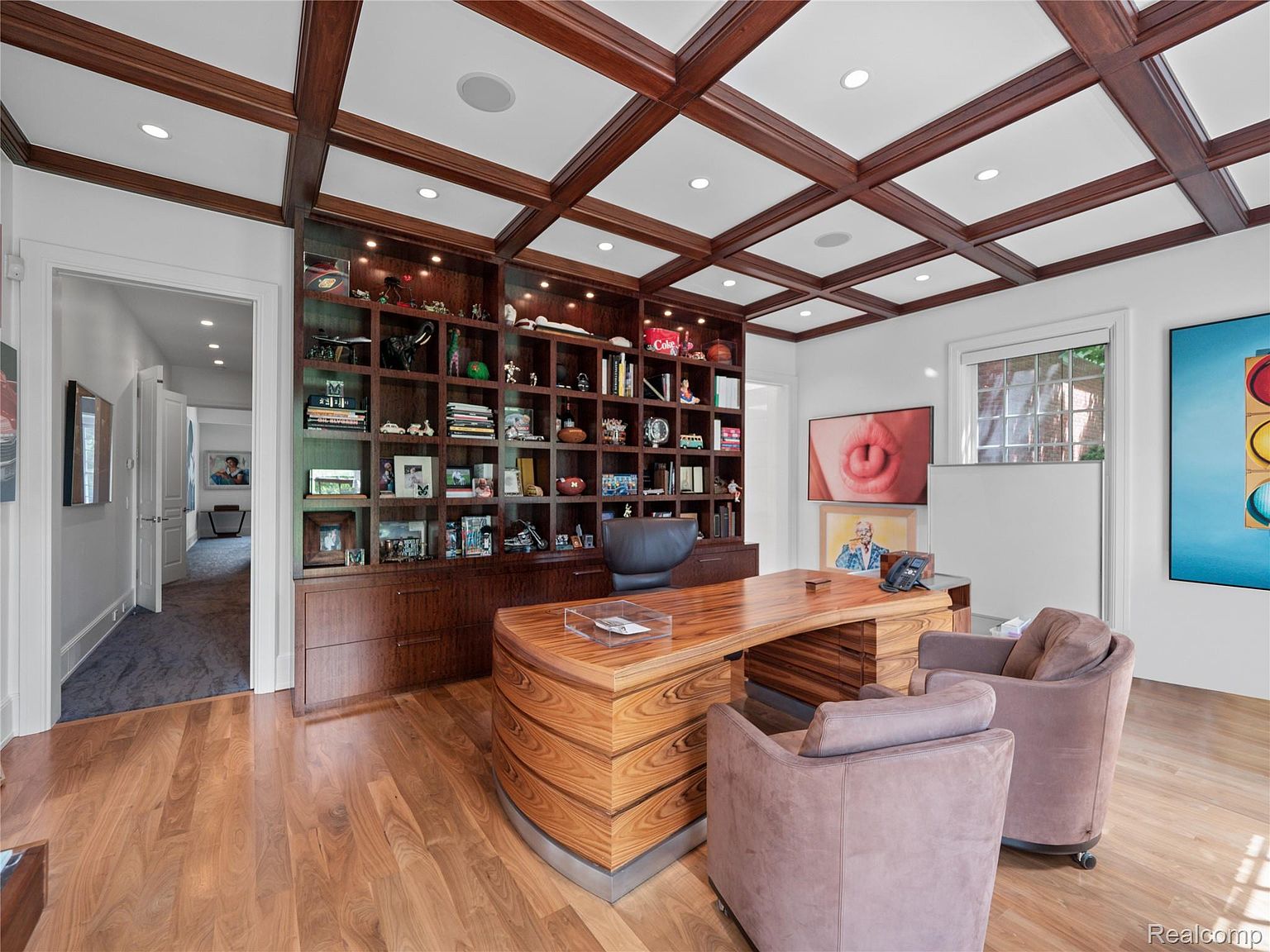 This is an interior shot of a well-appointed home office. The room features a custom wood coffered ceiling, a large built-in bookcase filled with decorative items, and a unique curved wooden desk. Two comfortable armchairs face the desk, and artwork adorns the walls, creating a sophisticated and inviting workspace.