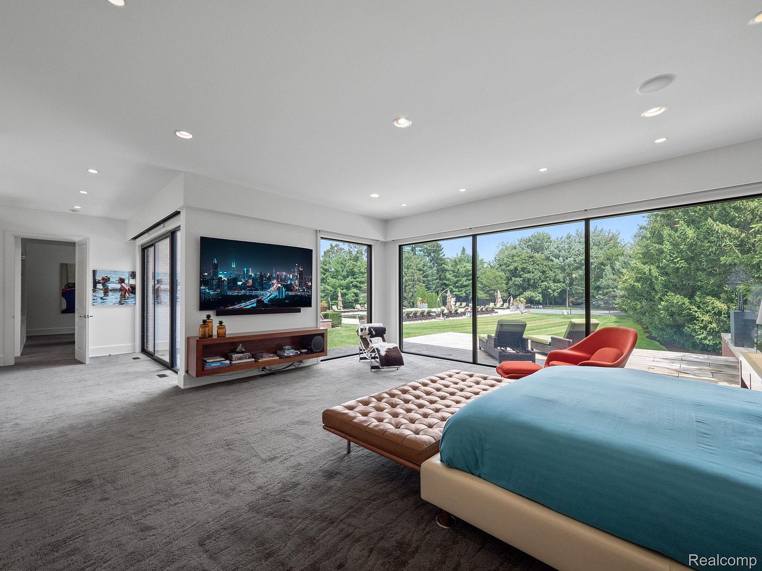 This is a luxurious primary bedroom featuring a large bed with a blue comforter and a tufted leather bench. The room has floor-to-ceiling windows offering a view of the expansive backyard. A modern entertainment center with a large screen TV and sleek shelving adds to the room's contemporary style.