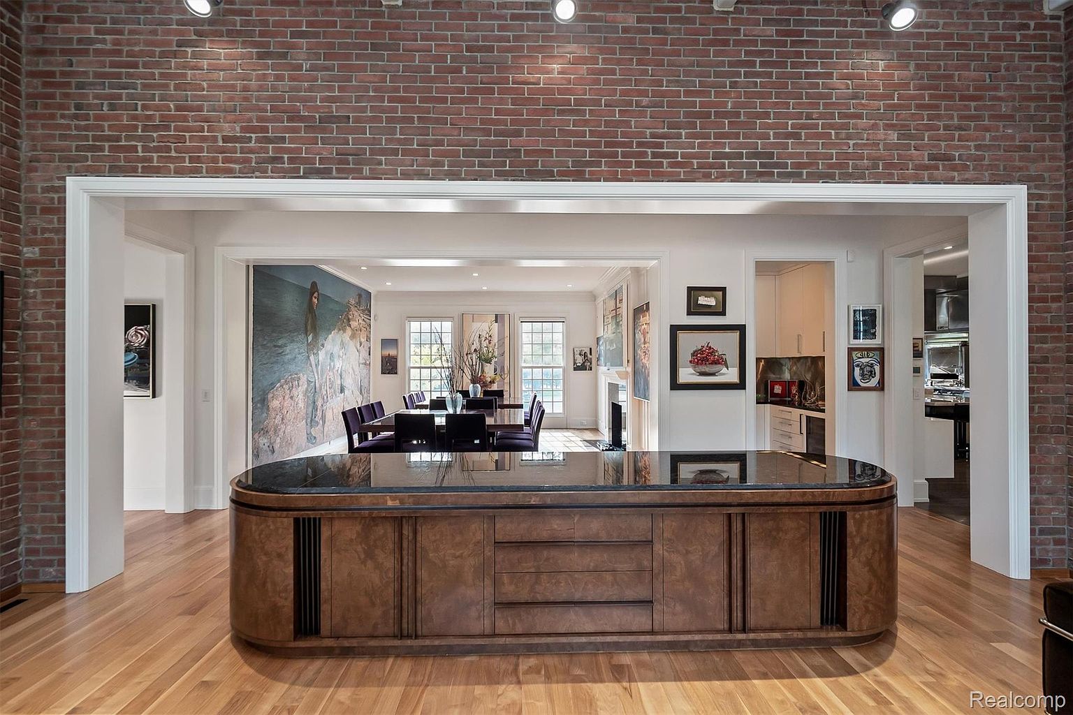 This interior shot showcases a dining room with a sophisticated design. The room features a large dining table with purple chairs, visible through a wide doorway framed by brick and white trim. A prominent island with a dark countertop and wood cabinetry sits in the foreground, adding depth and texture to the space.