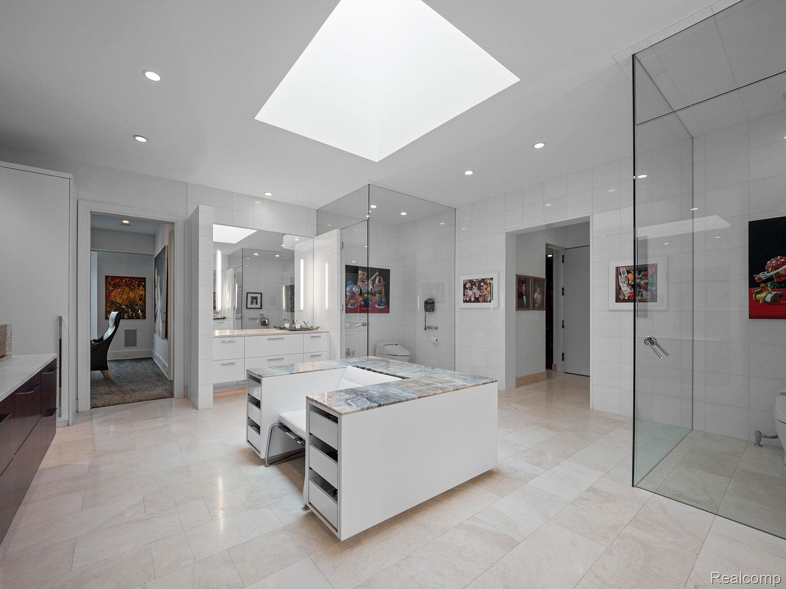 This is a spacious primary bathroom featuring a modern design with white tile walls and light-colored flooring. The bathroom includes a large skylight, a glass-enclosed shower, and a double vanity with marble countertops. There are also two islands with seating and storage, adding to the luxurious feel of the space.