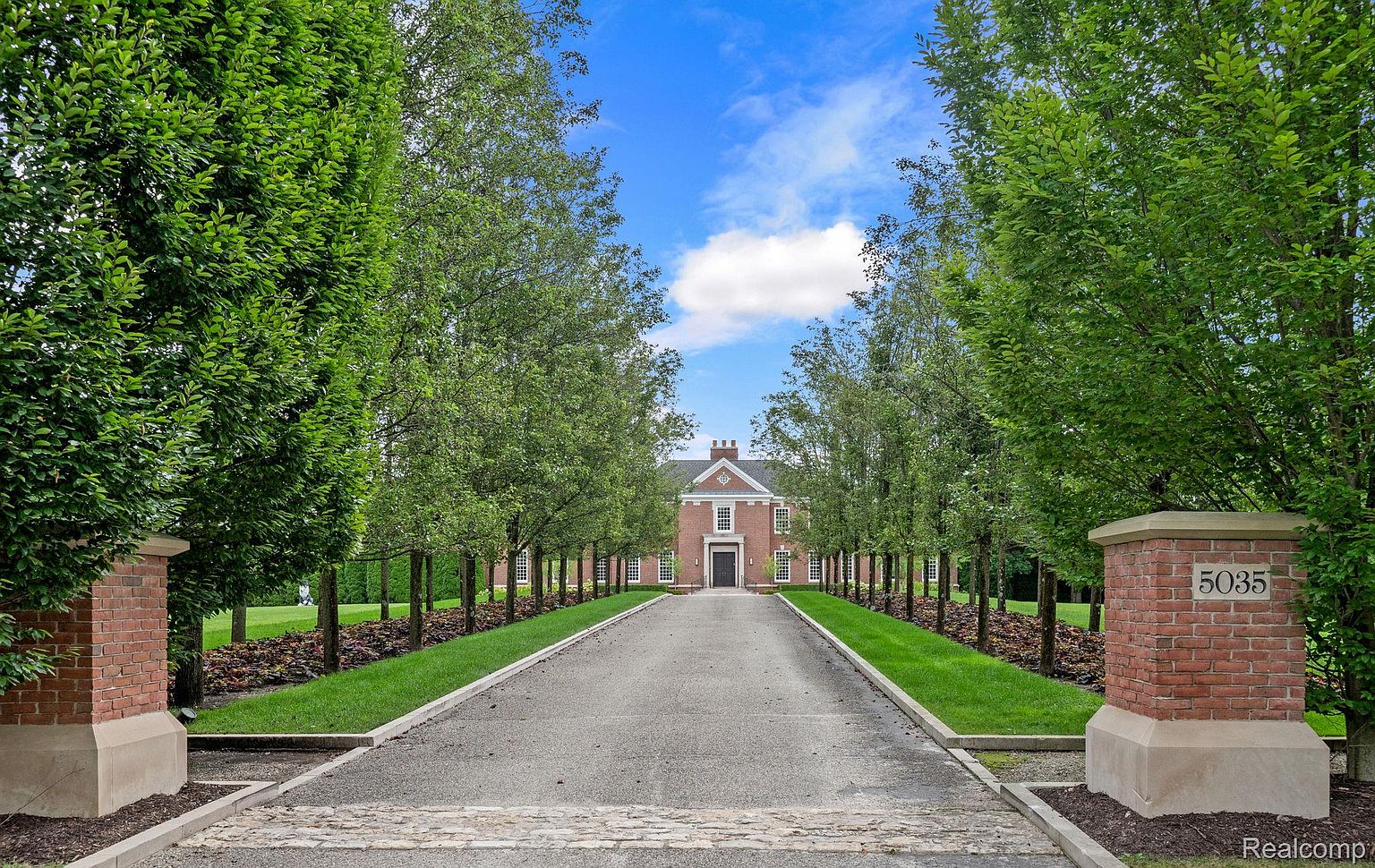 This is a front view of a stately brick home, showcasing a long, tree-lined driveway leading to the entrance. The symmetrical facade, manicured lawn, and brick pillars at the driveway entrance create a sense of grandeur and curb appeal. The house is framed by lush greenery and a clear blue sky, emphasizing its well-maintained and inviting appearance.