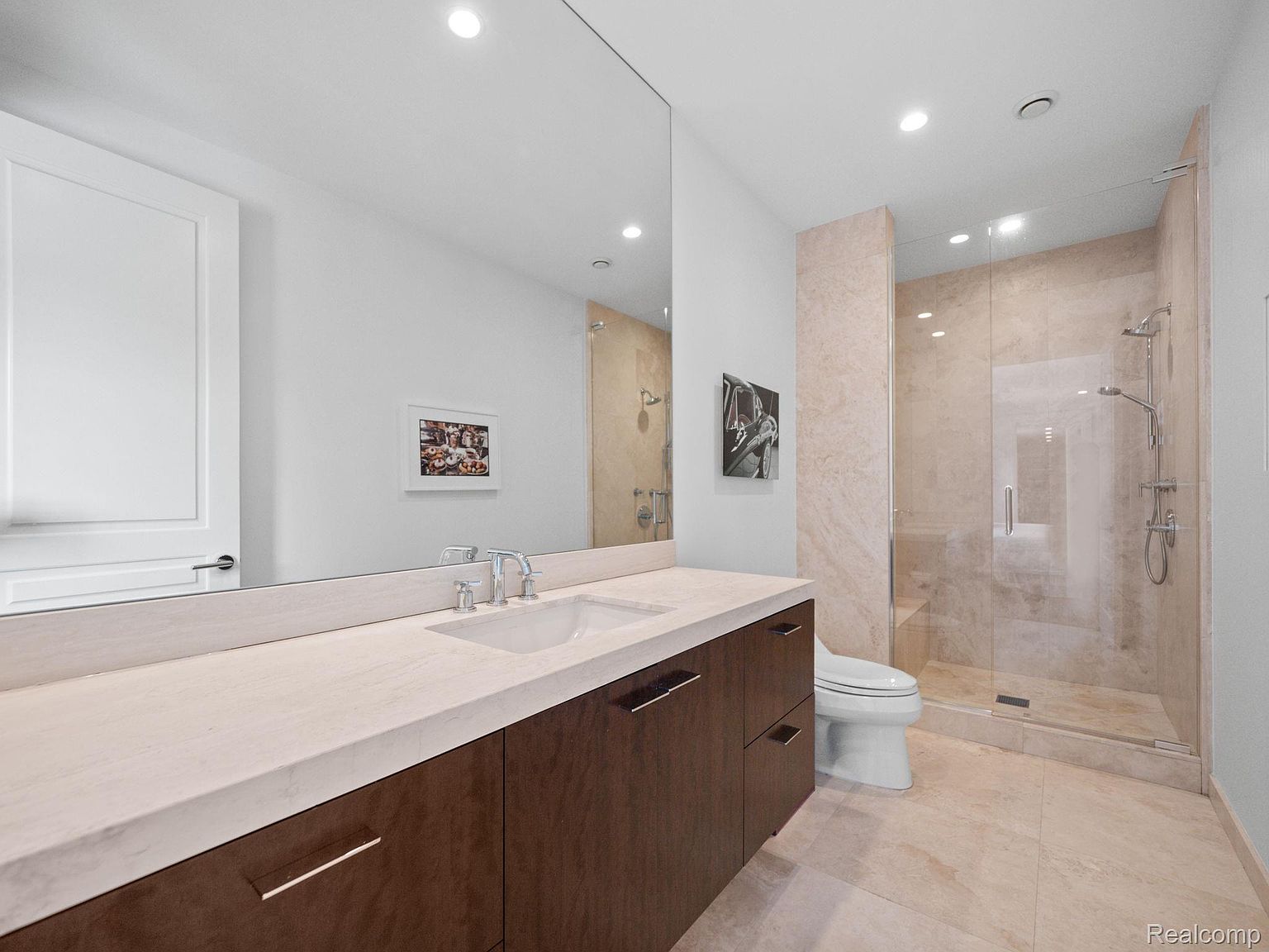 This is a primary bathroom featuring a modern design with a large mirror above a vanity with a light-colored countertop and dark wood cabinets. A glass-enclosed shower is visible to the right, with beige tiling and a rainfall showerhead. The overall impression is clean, luxurious, and well-lit.