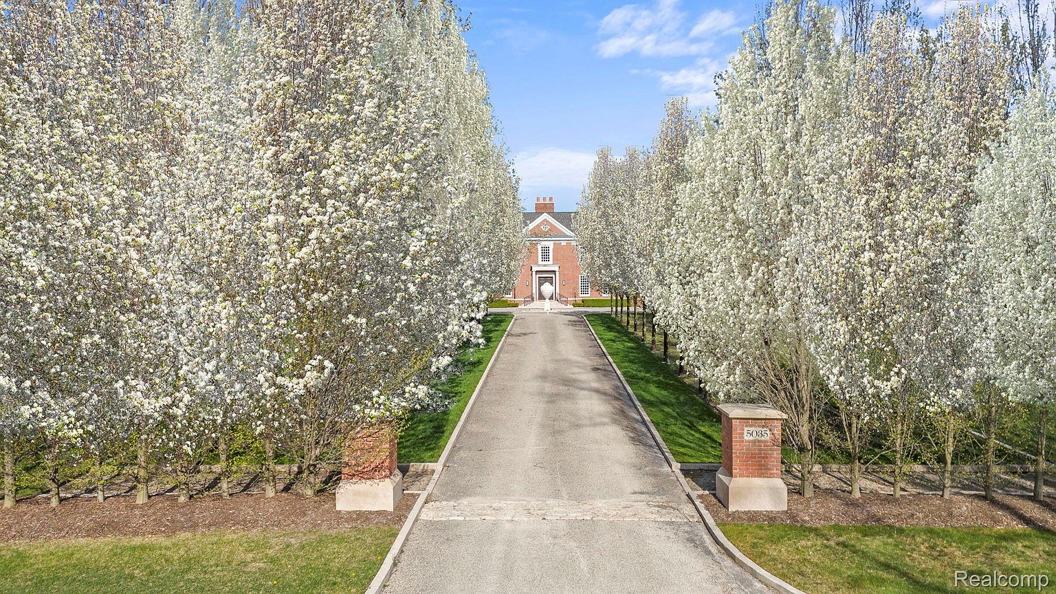 This is a captivating front view of a stately brick home, framed by a long driveway lined with blossoming trees. The symmetrical design of the house, coupled with the manicured lawn and mature landscaping, creates a grand and inviting entrance. A brick pillar with the house number is visible near the driveway entrance.