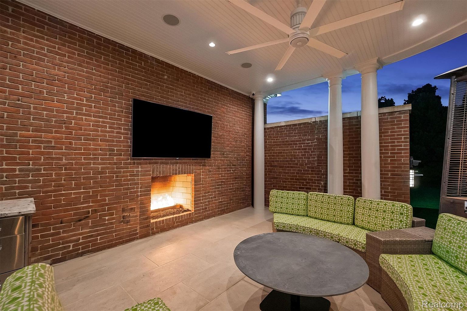 This is a well-appointed outdoor patio area featuring a brick wall with a fireplace and a mounted television. The space is furnished with comfortable seating around a round table, and the patio is covered with a ceiling fan and recessed lighting. The overall impression is one of a cozy and luxurious outdoor living space.