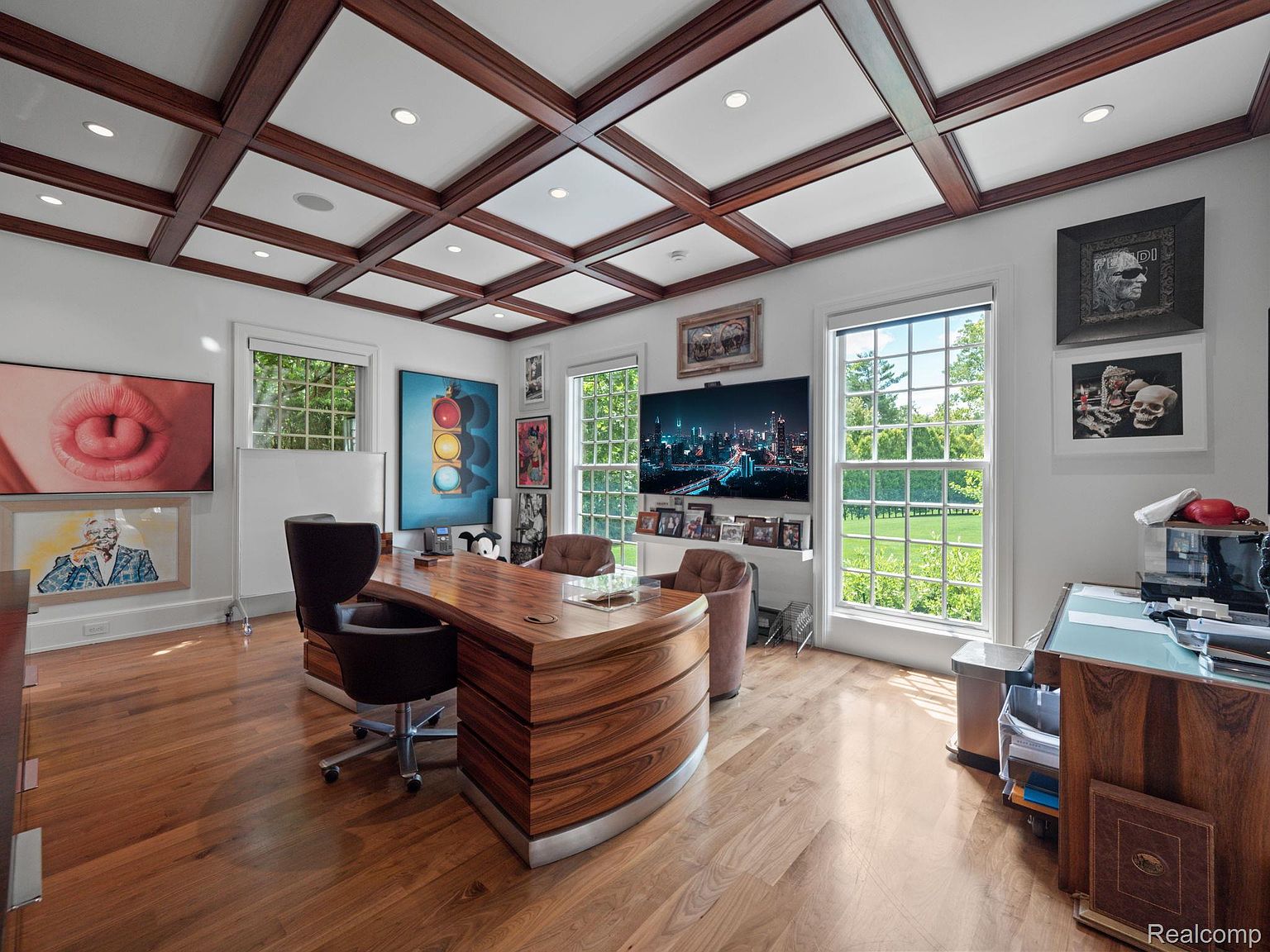 This is an interior shot of a well-appointed office space. The room features a unique, curved wooden desk as the focal point, complemented by leather chairs. Large windows provide ample natural light and views of the exterior, while artwork adorns the walls, adding character to the space. The coffered ceiling adds a touch of elegance.