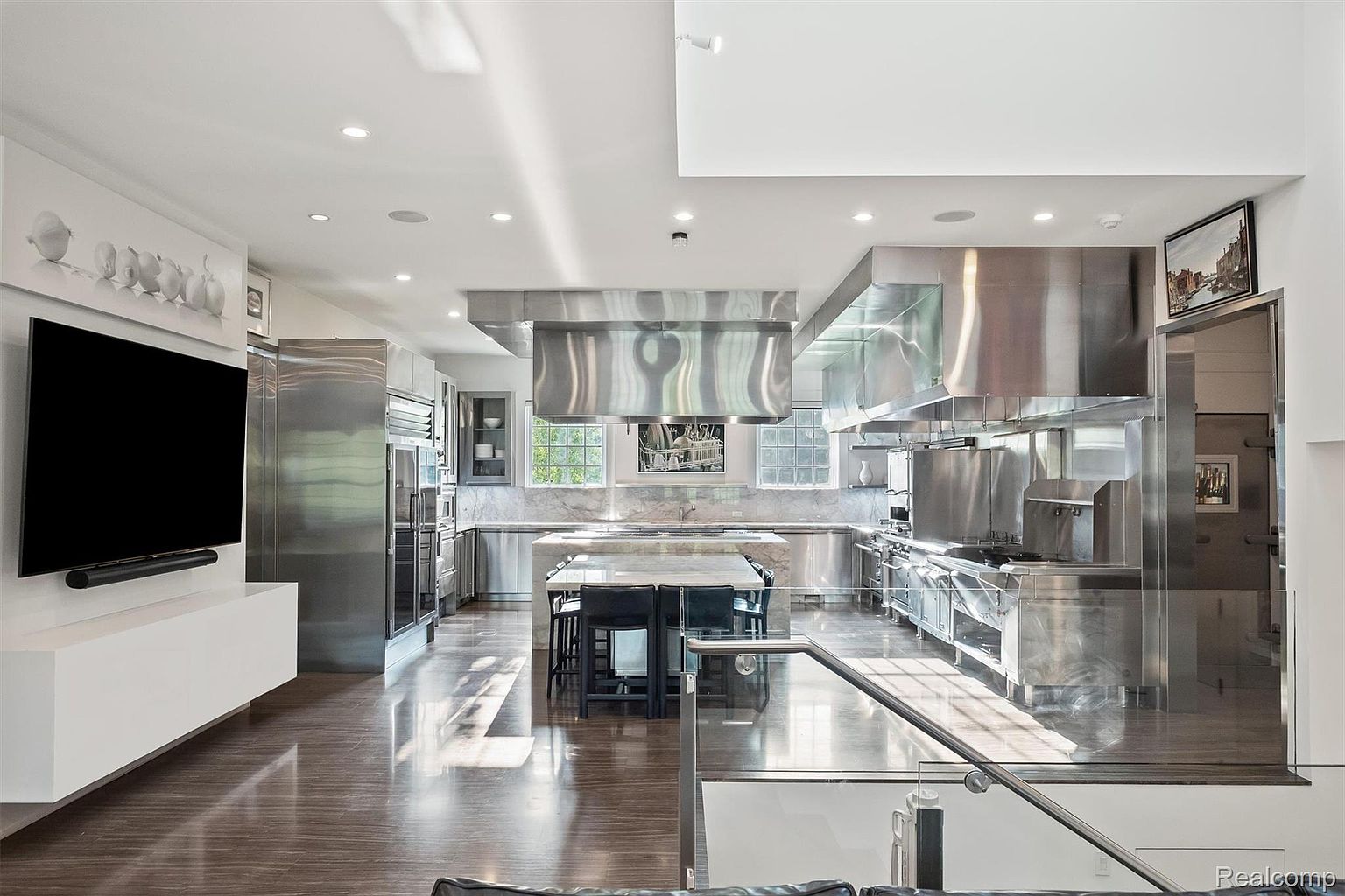 This is a spacious, modern kitchen featuring stainless steel appliances and cabinetry, a large central island with seating, and hardwood flooring. The kitchen is well-lit with natural light streaming through the windows, and the overall design is sleek and professional. A television is mounted on the wall in the adjacent living area.