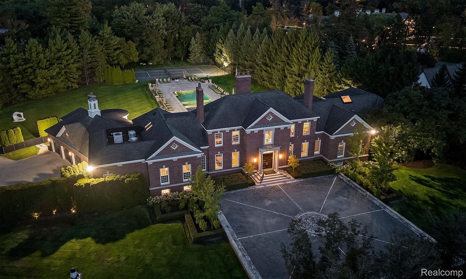 This aerial view showcases a grand estate with a brick facade, dark roof, and multiple chimneys. The property features a circular driveway, manicured lawns, a swimming pool, and a tennis court, all surrounded by mature trees. The overall impression is one of luxury and privacy.