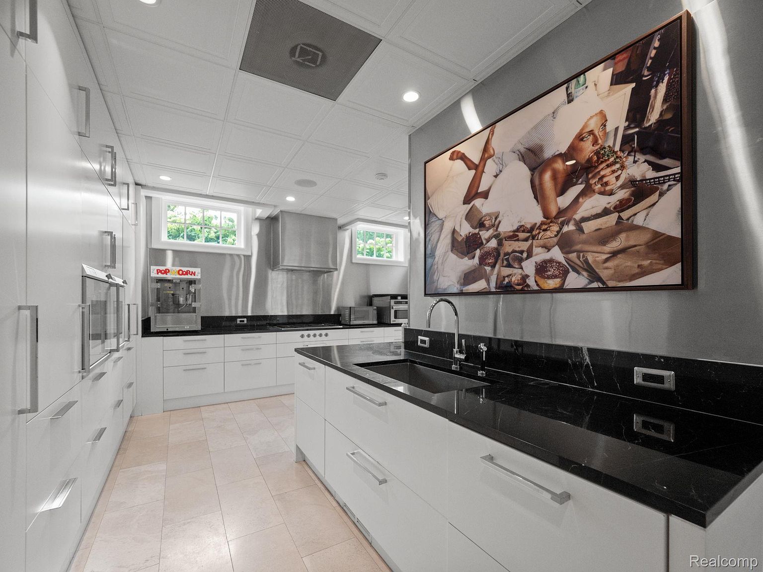 This is a modern kitchen with white cabinetry, black countertops, and stainless steel appliances. The kitchen features a large piece of art on the wall, a popcorn machine, and a unique ceiling design. The overall impression is clean, bright, and stylish.