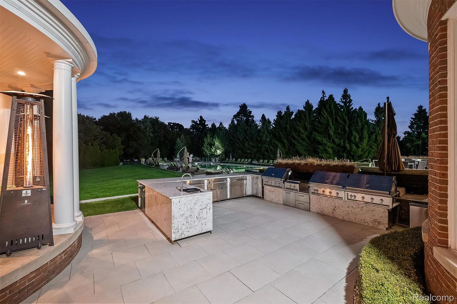 This image showcases a luxurious outdoor kitchen and patio area at dusk. The space features high-end stainless steel grills and appliances built into a stone countertop, along with a large island. The patio is paved with large stone tiles, and the area is surrounded by manicured landscaping and mature trees, creating a private and upscale outdoor living space.