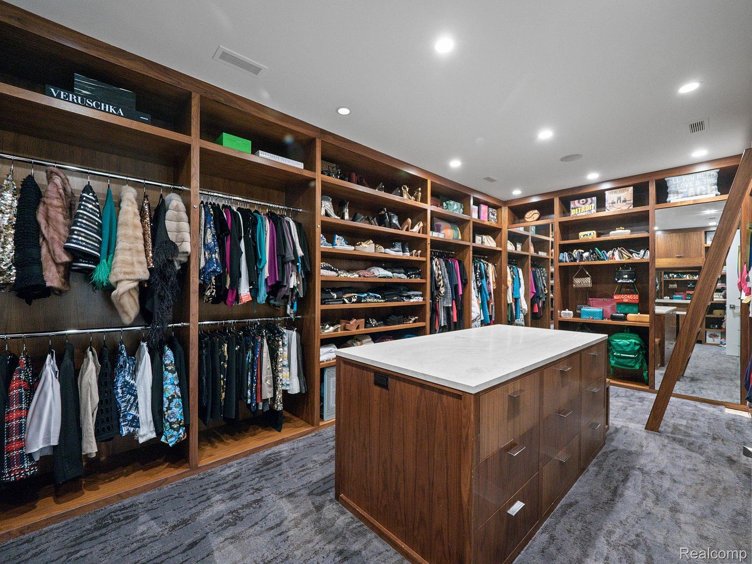 This is a spacious walk-in closet featuring custom wood shelving and hanging rods filled with clothing and accessories. A central island with a white countertop and drawers provides additional storage and workspace. The closet is well-lit with recessed lighting, and the overall impression is luxurious and organized.