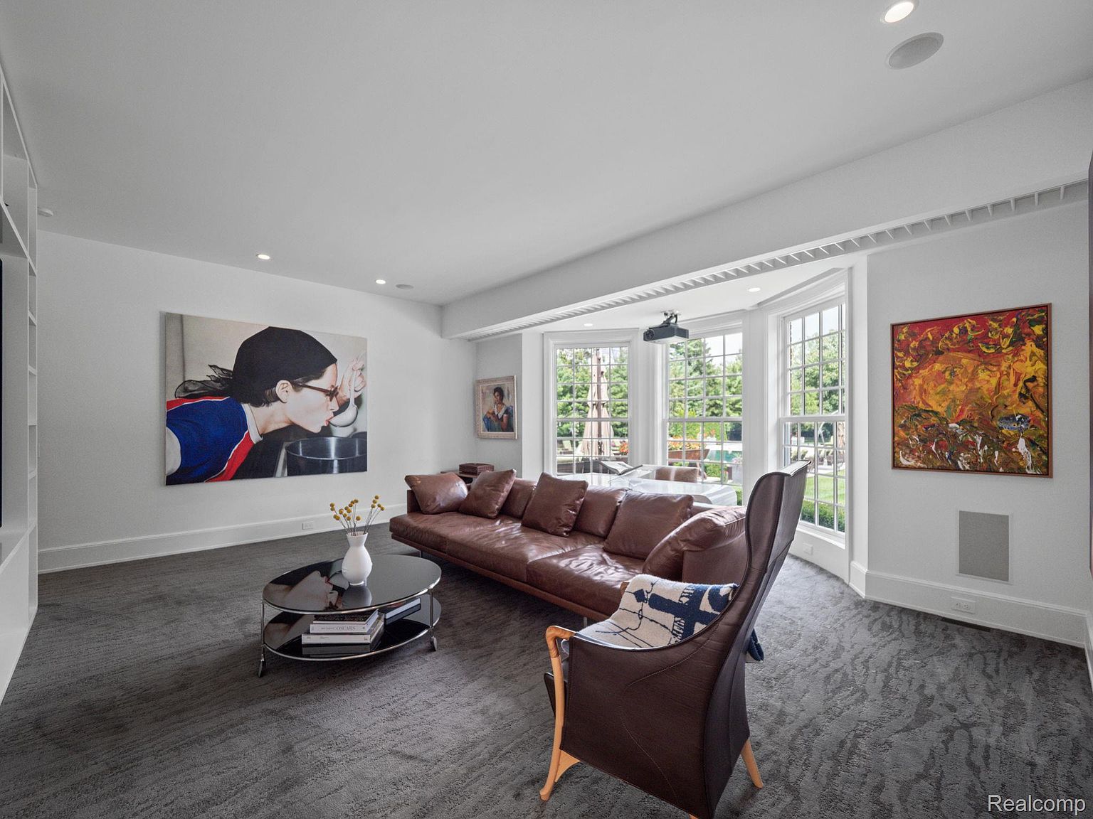 This is an interior shot of a living room featuring a brown leather sofa, a brown leather chair, and a black glass coffee table. The room has large windows offering natural light and a view of the outdoors. Artwork adorns the walls, and the overall aesthetic is modern and comfortable.