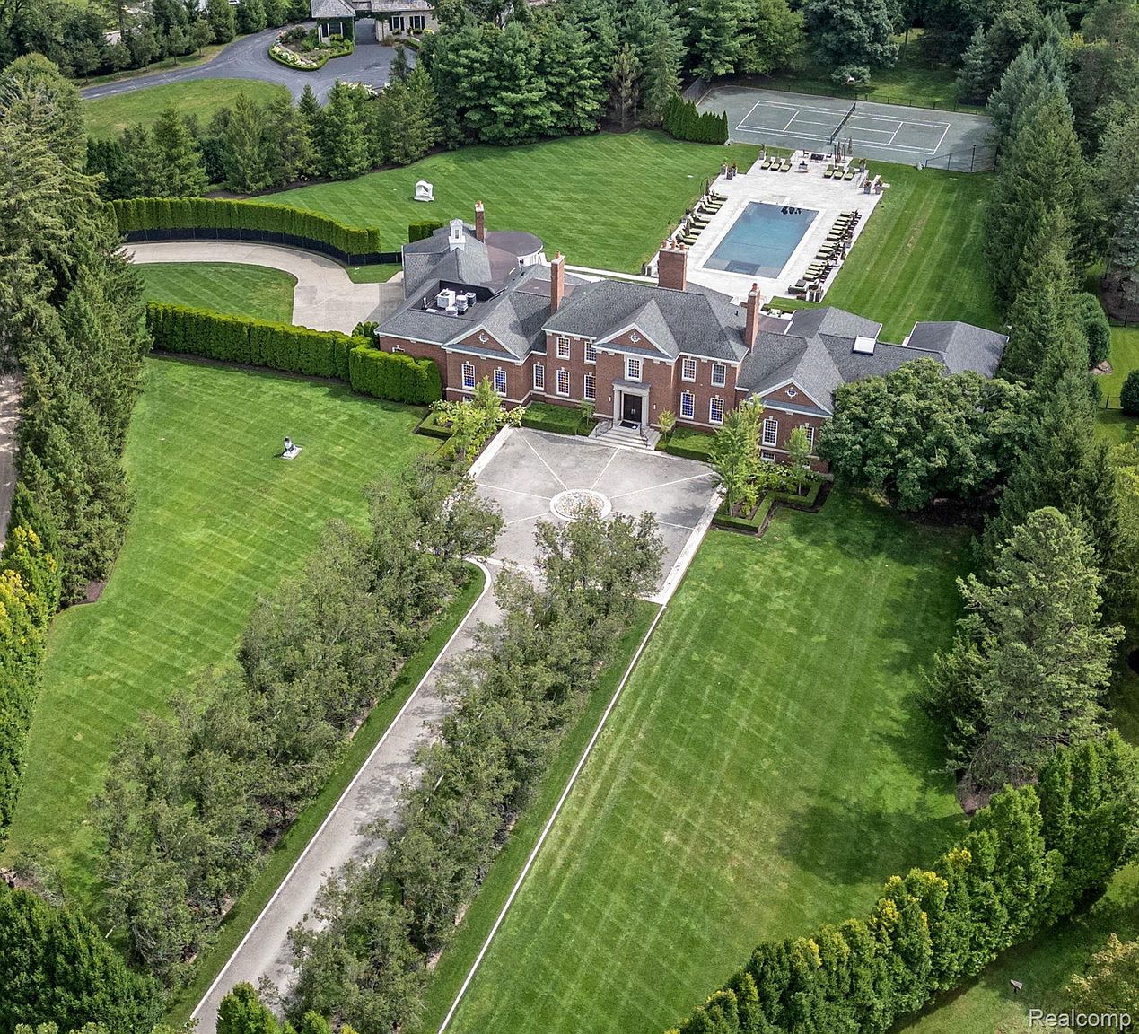This aerial shot showcases a grand estate with meticulously manicured lawns, mature trees, and formal gardens. The brick mansion features a symmetrical facade, a circular driveway, and a pool with a tennis court in the backyard. The overall impression is one of luxury and privacy.