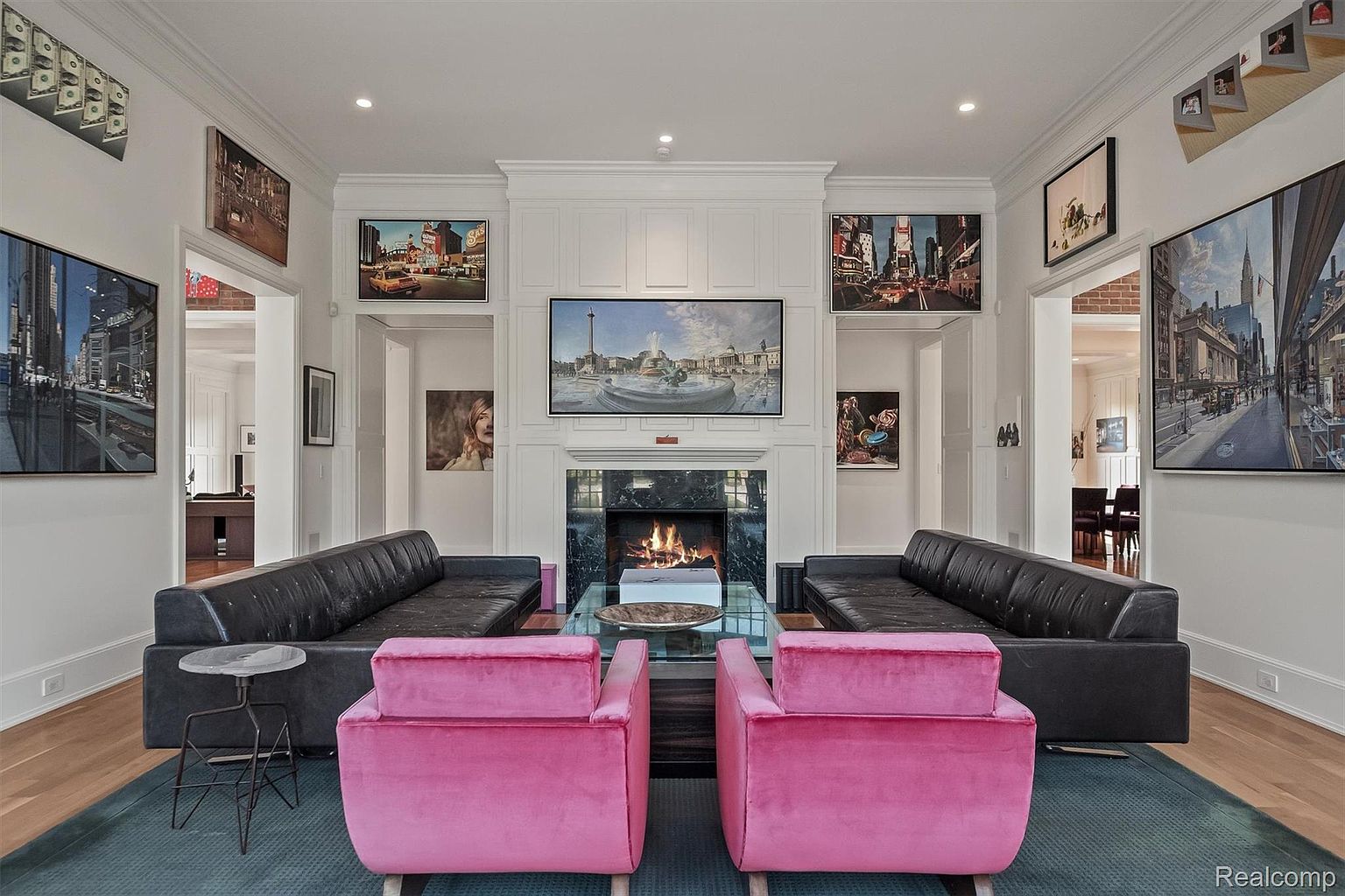 This is an interior shot of a luxurious living room featuring two black leather sofas facing each other, with two pink velvet armchairs in the foreground. A glass coffee table sits in the center, and a fireplace with a marble surround is the focal point. Artwork adorns the walls, and the room is flanked by doorways leading to other parts of the house, creating a sense of spaciousness and elegance.